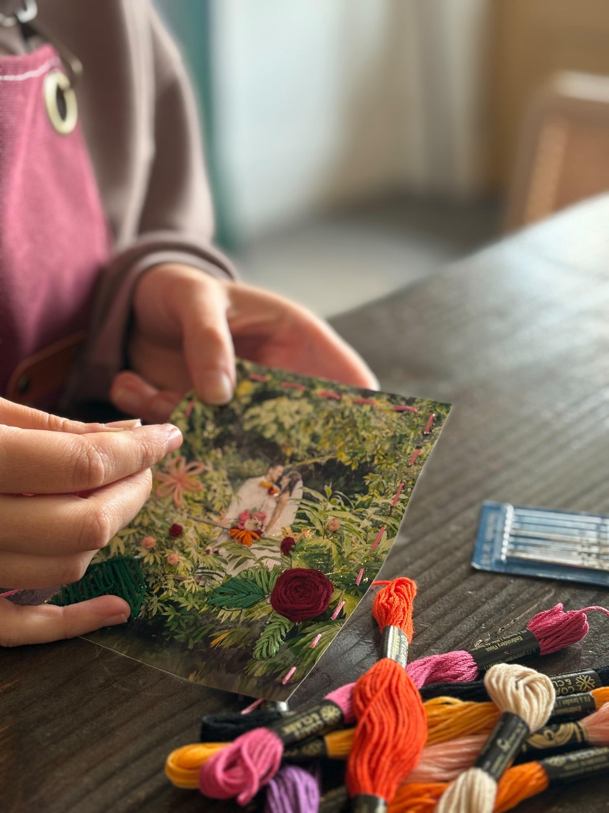 Child's hands holding an embroidery pattern with colorful floss skeins arranged on a table.