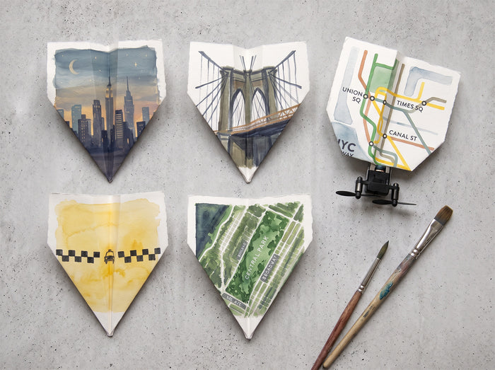 Five paper airplanes decorated with NYC landmarks and a subway map, with paintbrushes on a concrete surface.