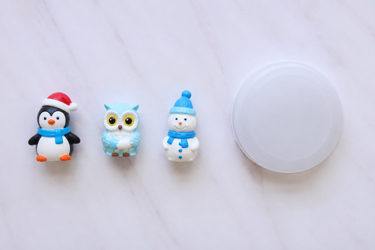 Painted penguin, owl and snowman figures lined up beside a white snow globe base