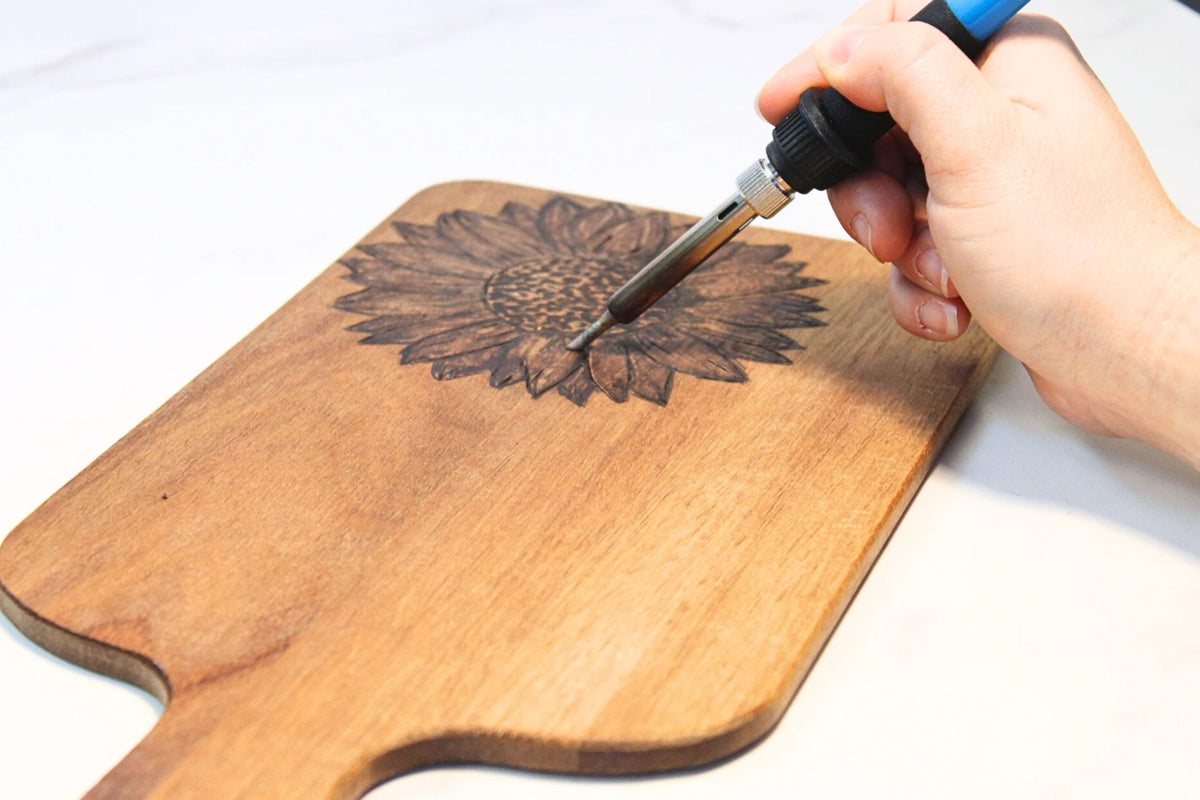 Hand guiding a wood burning pen along a sunflower design on a wooden board