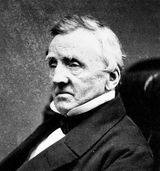 Close-up black and white portrait of a man in a suit, highlighting his face and attire, suitable for a professional or formal craft workshop setting.