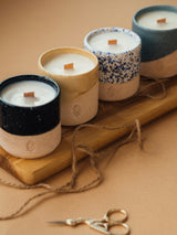 A group of candles, including one in a blue and white container and one with a wooden stick, displayed on a wooden board.