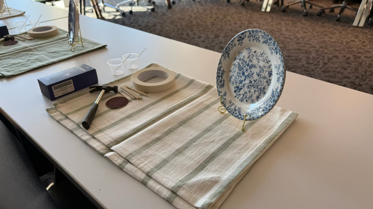 A striped linen placemat displays ceramic plates, utensils, and a decorative blue-and-white plate on an easel.