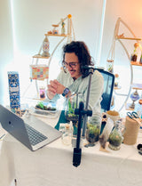 A creative individual with long hair and glasses beams while seated at a desk adorned with an array of plants and containers, checking their smartwatch. As the host for engaging virtual team-building craft workshops, a laptop and microphone stand take their place on the crafting table. Shelves filled with crafting supplies decorate the background, setting the perfect scene for an inspiring experience.