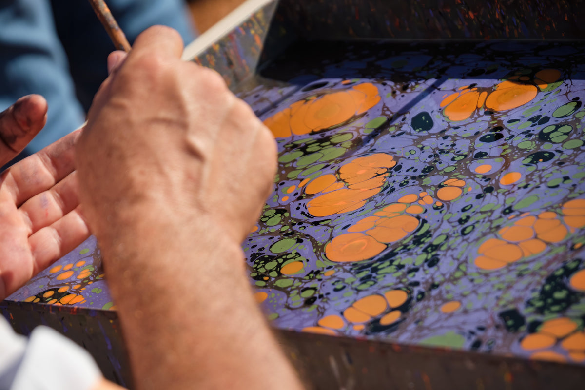 Hands raking floating pigment into a marbled Ebru pattern on a tray of water