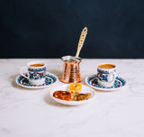 Turkish Coffee Workshop, Two beautifully decorated coffee cups with matching saucers, a charming copper pot featuring a long handle, and an artfully arranged plate of assorted snacks rest gracefully on a marble surface. The dark background provides an elegant contrast, highlighting the intricate details of this sophisticated setup.