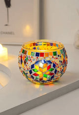 A vibrant mosaic-patterned candle holder rests on a white surface, radiating a warm, glowing light from the candle it cradles within.