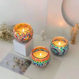 Three illuminated mosaic glass candle holders in assorted sizes grace a pristine white table. Surrounding them are charming decorative accents like a faceless wooden mannequin and dried floral arrangements, adding an artistic touch to the scene.