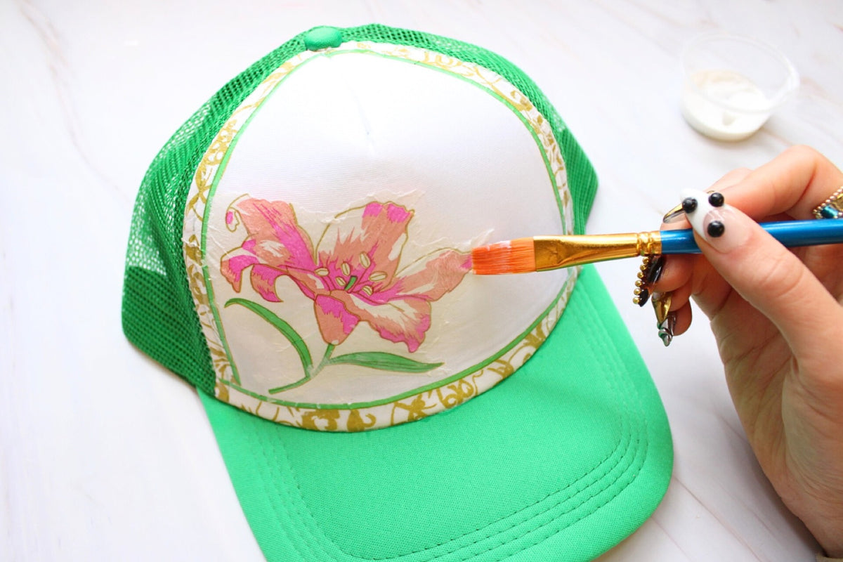 Hand painting a pink flower design onto the front of a green trucker hat