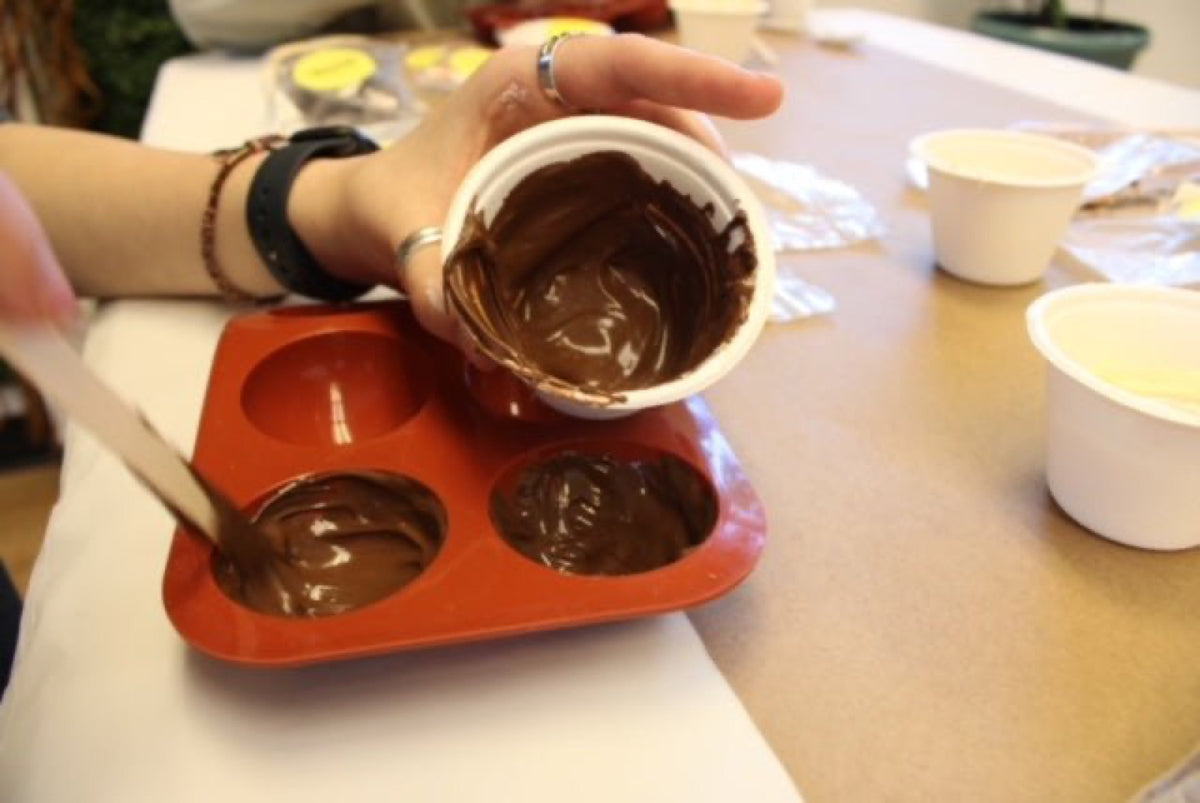 Hands pouring melted chocolate from a jug into a red silicone bomb mould