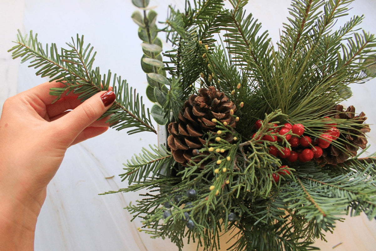 Hands layering evergreen and berry sprigs onto a wreath